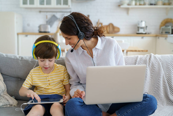 Šťastná matka s dítětem sedí na pohovce doma během uzamykání práce na notebook dítě hraje v tabletu - Fotografie, Obrázek