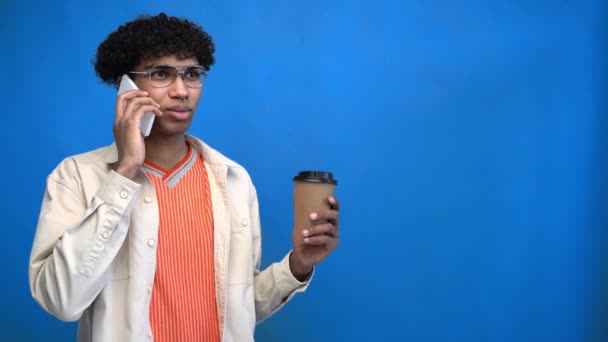 Afrikaan amerikaanse man met papieren beker en praten op mobiele telefoon geïsoleerd op blauw - Video