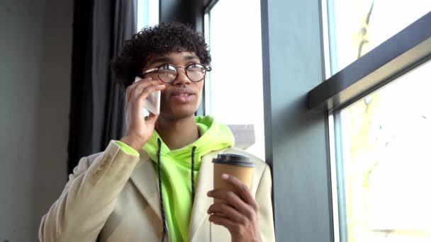 joven hombre de negocios afroamericano hablando en el teléfono inteligente y sosteniendo taza de papel - Metraje, vídeo