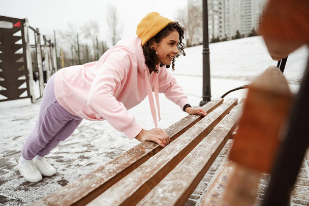 smiling african american sportswoman doing push ups near bench outdoors, blurred foreground - Photo, Image