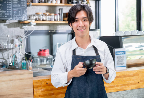 コーヒーメーカーのバリスタの男がカップを持ち、カメラと笑顔でコーヒーショップのカウンターの前に立っている。中小企業との幸せな作業の概念と持続可能な. - 写真・画像