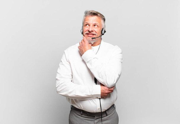 senior telemarketer smiling with a happy, confident expression with hand on chin, wondering and looking to the side - Photo, Image