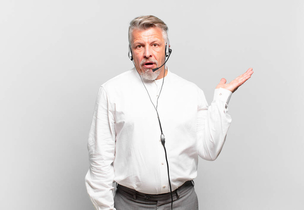 senior telemarketer looking surprised and shocked, with jaw dropped holding an object with an open hand on the side - Foto, Imagem