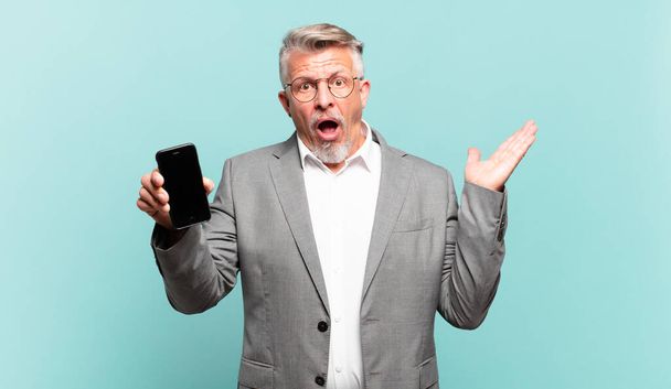 senior businessman looking surprised and shocked, with jaw dropped holding an object with an open hand on the side - Foto, afbeelding