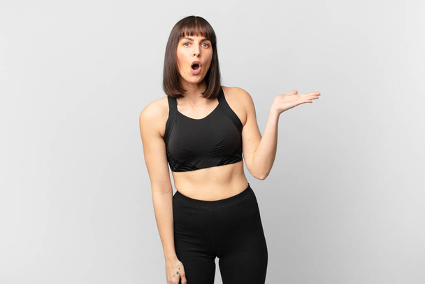 athlete woman looking surprised and shocked, with jaw dropped holding an object with an open hand on the side - Foto, afbeelding