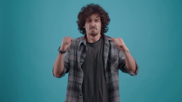 Nervous hipster with curly hair, shouts, disappointed puts his hands on his head. Young man dressed in a plaid shirt, on a blue background, in the studio. - Footage, Video