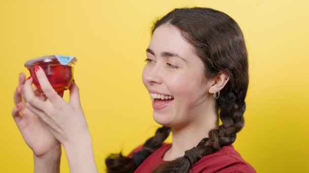 Een jonge glimlachende vrouw opent een pot gelei, ruikt eraan en ruikt een vreselijke geur. Gele achtergrond. Het concept van junkfood. - Video