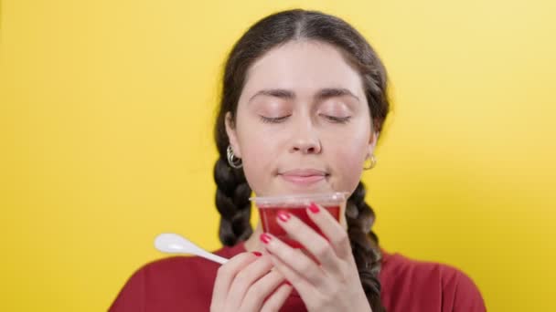 Portrait d'une jeune femme souriante qui renifle un pot parfumé de gelée, et met soigneusement une cuillère à sa bouche. Fond jaune. Concept de régime alimentaire. - Séquence, vidéo