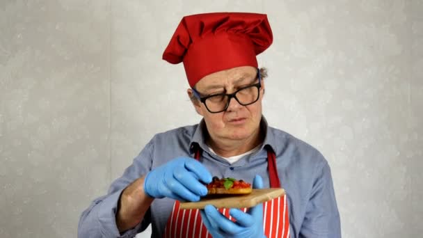 Senior volwassen chef-kok Kaukasische etniciteit in rode chef-kok hoed, proeven met genieten van bruschetta met tomaten en basilicum, in de binnenlandse keuken. Close-up. - Video