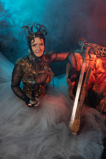 Fabulous creature, queen, sorceress, in a colorful suit and sword. Woman, model after 60, in the studio, dark background - Photo, Image