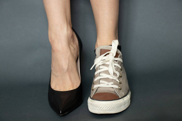 на одной ноге черные туфли на высоком каблуке на другой спортивные кроссовки на ногах девушки на изолированном темно-синем фоне - Фото, изображение