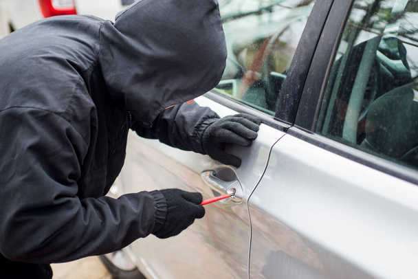 Robber man in black hoodie jacket using a screwdriver  to break lock and steal a vehicle. Car thief or theft for insurance concept. - Photo, Image