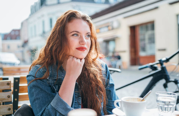 Portrait of sad red curled long hair caucasian woman sitting on a cozy outdoor cafe terrace on the street and looking at the street. Young woman taking a break in her city bicycle tour. - Φωτογραφία, εικόνα