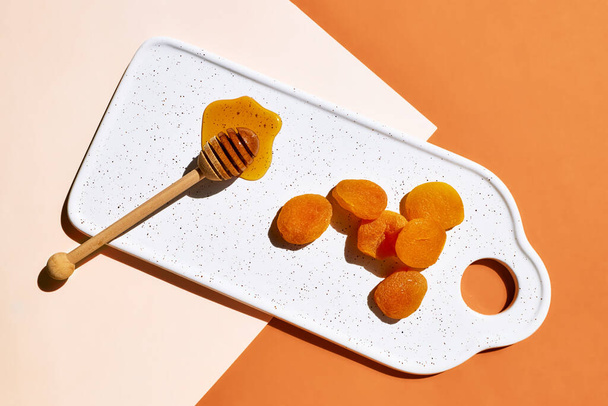 Сушені абрикоси та мед у білій тарілці на помаранчевому фоні. Зневоднені плоди. Концепція здорових цукерок. Вид зверху
 - Фото, зображення