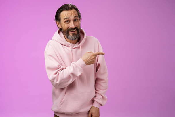 Barátságos gondtalan kaukázusi szakállas férfi rózsaszín kapucnis mutatva jobbra intoducing családtagok mosolyogva büszkén mutatja üres másolás tér reklám, pózol lila háttér - Fotó, kép