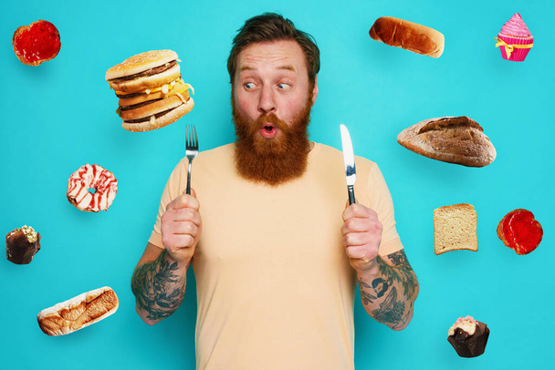 Der tätowierte Mann ist bereit, mit Besteck in der Hand Sandwich und Süßigkeiten zu essen. Cyan-Hintergrund - Foto, Bild