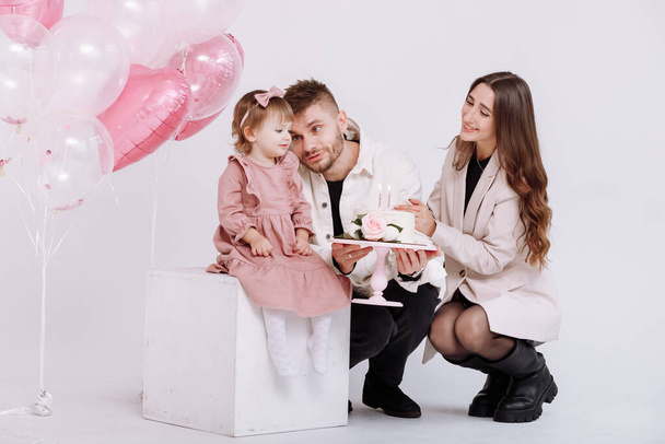 Mladí rodiče oslavují narozeniny svého dvouletého dítěte. rodinné světlo a sfouknutí svíčky na dort s květinami na pozadí narozeninové výzdoby s růžovými kuličkami - Fotografie, Obrázek