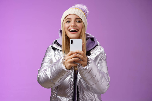 Διασκέδαση ενθουσιασμένοι ελκυστική ξανθιά φίλη κρατώντας smartphone μέχρι την εγγραφή βίντεο φίλο βήμα snowboard πρώτη φορά συλλαμβάνοντας τις αναμνήσεις κινητή κάμερα στέκεται ευτυχώς μωβ φόντο - Φωτογραφία, εικόνα