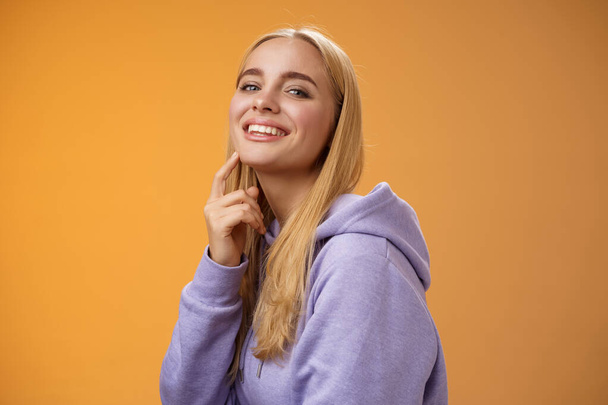 Attraktive freche selbstbewusste kaukasische Blondine in lila Kapuzenpulli, die Kamera anrührt Kinn frech lächeln Kamera schaut interessante Sache fasziniert nach vorne, stehend orangefarbenen Hintergrund - Foto, Bild