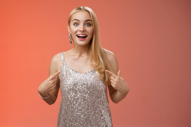 Удивлена удивлена счастливая блондинка очаровательная веселая женщина в блестящем серебряном платье указывая себя забавно взволнован выбран участвовать удивительное событие, стоя радостный красный фон - Фото, изображение