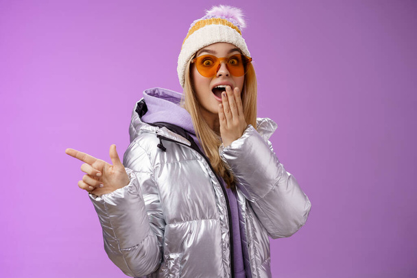 Amazed speechless surprised attractive stylish blond girlfriend in sunglasses, silver glittering jacket hat drop jaw impressed widen eyes shocked pointing left astonished, purple background - Photo, Image