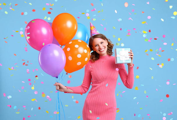 Renkli balonlar ve hediye kutusuyla iyimser bir kadın düşen konfeti altında duruyor ve doğum günü kutlaması sırasında mavi arka planda kameraya gülümsüyor. - Fotoğraf, Görsel