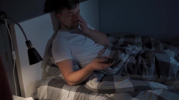 Zmęczony człowiek trzyma pilota w rękach i zasypiając, ogląda telewizję w nocy w łóżku. Tematyczna zależność od usług streamingowych, filmów i programów telewizyjnych. Mężczyzna cierpi na bezsenność w nocy - Materiał filmowy, wideo