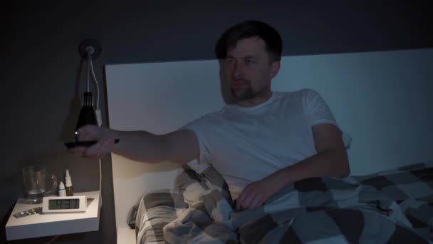Young caucasian man suffering from depression and insomnia getting upset trying to entertain himself while watching tv via streaming service, switching channels with remote control in bed at night - Materiaali, video