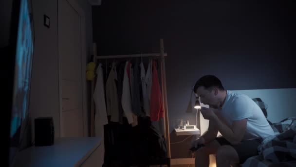 Młody mężczyzna rasy kaukaskiej cierpiący na depresję i bezsenność denerwuje się próbując zabawić się podczas oglądania telewizji za pośrednictwem serwisu streamingowego, przełączając kanały z pilotem w łóżku w nocy - Materiał filmowy, wideo