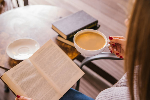 Mladá dívka sedí v kavárně a čte si knihu. Ta holka pije kafe nebo kakao. Útulná atmosféra a příjemný volný čas. Odpočinek a relaxace. - Fotografie, Obrázek