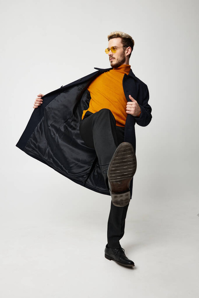 fashionable man in a coat and trousers is dancing on a light background and an orange sweater glasses - Фото, изображение