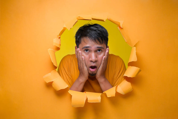 Frustrovaný Asiat pózuje roztrhanou žlutou dírou v papíře, bojí se, zmatený muž v brýlích ohromený nečekanými zprávami nebo informacemi - Fotografie, Obrázek