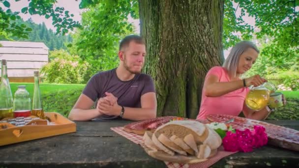 Guy hungry waiting patiently to start eating, Caucasian couple picnicking on a table under a tree, Dolly shot - Footage, Video