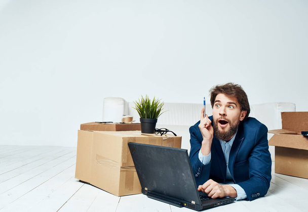 Un hombre con traje yace en el suelo con cajas de oficina desempacando la tecnología de un portátil - Foto, Imagen
