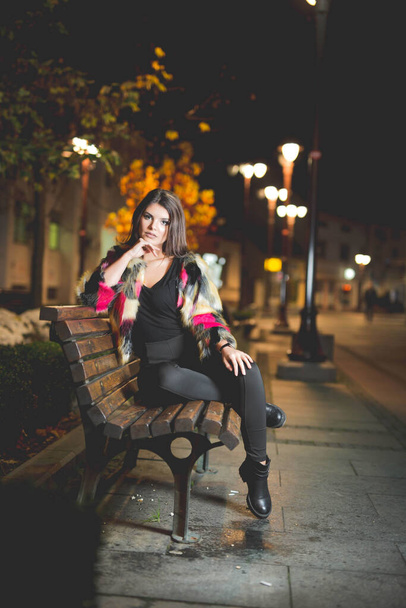 A vertical shot of a female in a fashionable jacket posing sitting on a bench in the street at night - Foto, Bild