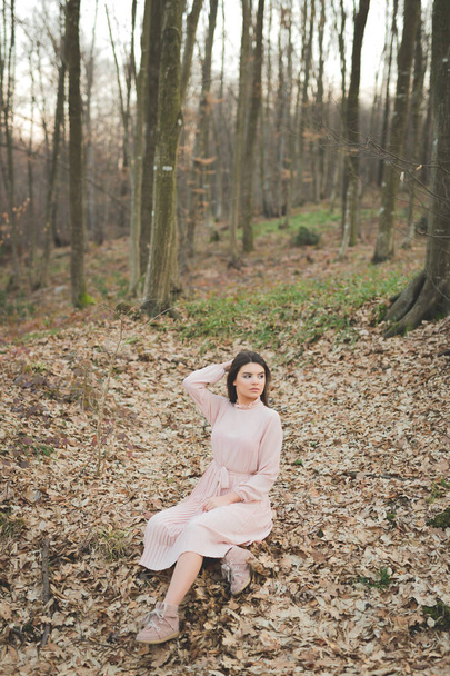 A young caucasian female in a delicate pink dress sitting on the fallen dry trees in a forest - Foto, afbeelding