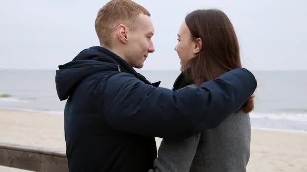 Soğuk havada deniz kenarında genç bir çift. Bir adam arka planda kız arkadaşını nazikçe öpüyor. Bir deniz var. - Video, Çekim
