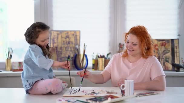 Madre e figlia dipingono insieme a casa con pennelli e acquerelli. Giovane donna seduta e aiutare o insegnare figlia a disegnare. 4k filmati - Filmati, video