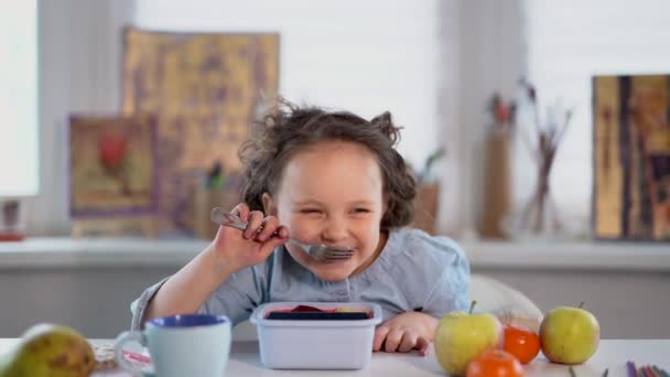Kisiskolás lány eszik, vagy ebédel. A lány villával eszi a kajáját és evés közben nevet. Boldog mosolygó kislány vacsorázik az asztalnál. 4k felvétel - Felvétel, videó
