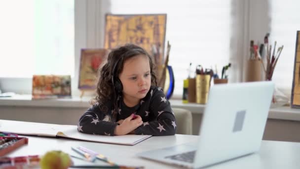 E-Learning-Stil für kleine Mädchen. Kind mit Kopfhörer und Laptop sitzt am Tisch und hört zu oder kommuniziert online. Online-Studium oder Lernkonzept. 4k Filmmaterial - Filmmaterial, Video