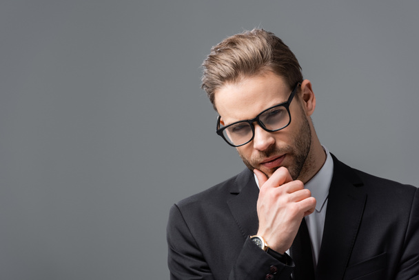 giovane uomo d'affari con gli occhiali che tocca il mento mentre pensa isolato sul grigio - Foto, immagini