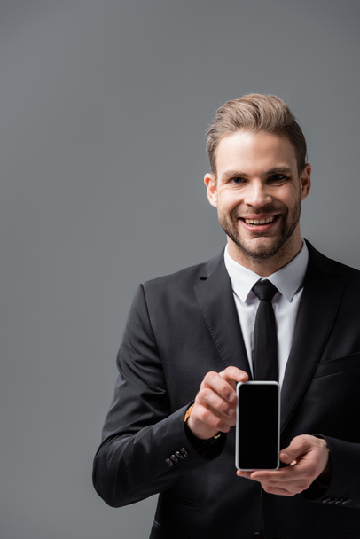 radosny biznesmen trzymający telefon komórkowy z pustym ekranem odizolowanym na szaro - Zdjęcie, obraz