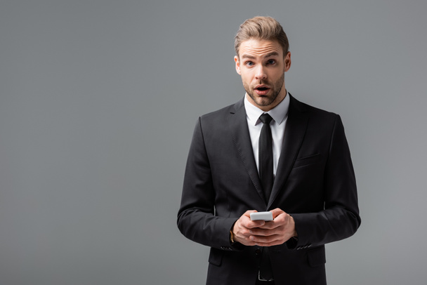 sorprendido hombre de negocios mirando a la cámara mientras charlaba en el teléfono inteligente aislado en gris - Foto, Imagen