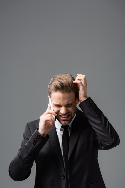 hombre de negocios estresado tocando la cabeza mientras habla en el teléfono inteligente aislado en gris - Foto, Imagen