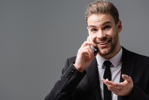 alegre hombre de negocios señalando con la mano mientras habla en el teléfono móvil aislado en gris - Foto, Imagen