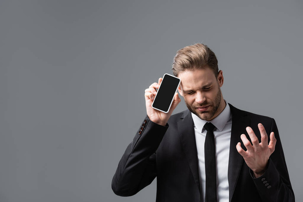 frustrovaný manažer gestikuluje při zobrazování mobilu s prázdnou obrazovkou izolovanou na šedé - Fotografie, Obrázek