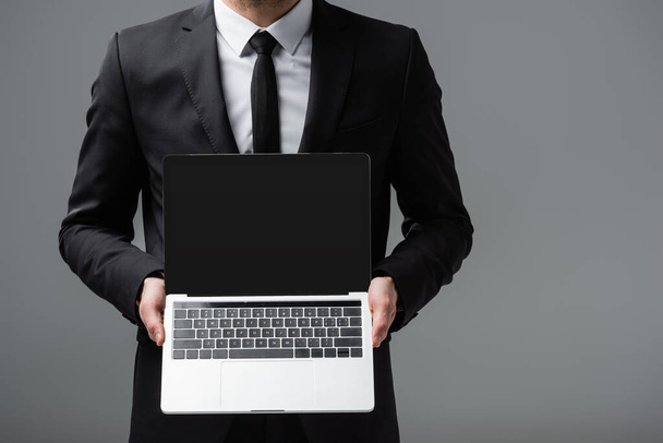 vista recortada del empresario que sostiene el ordenador portátil con pantalla en blanco aislado en gris - Foto, Imagen