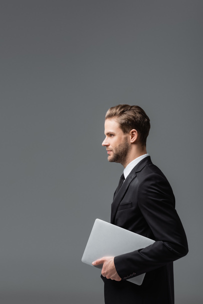 boční pohled na mladého výkonného manažera držícího notebook izolovaný na šedé - Fotografie, Obrázek