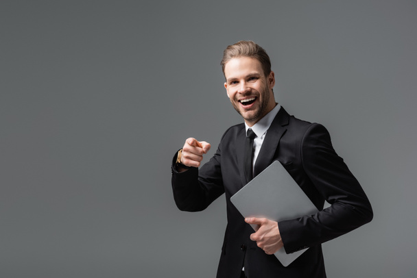 Смеющийся бизнесмен показывает пальцем, держа ноутбук изолированным на черном - Фото, изображение