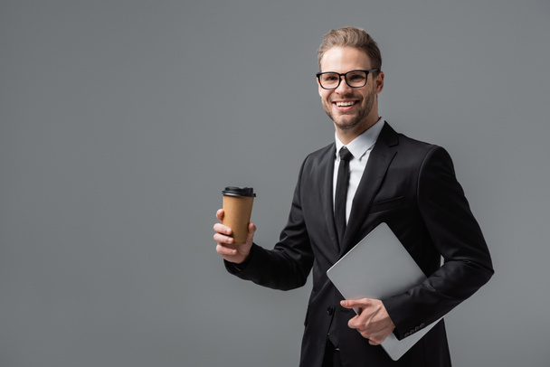 happy businessman with takeaway drink and laptop smiling at camera isolated on grey - Photo, Image